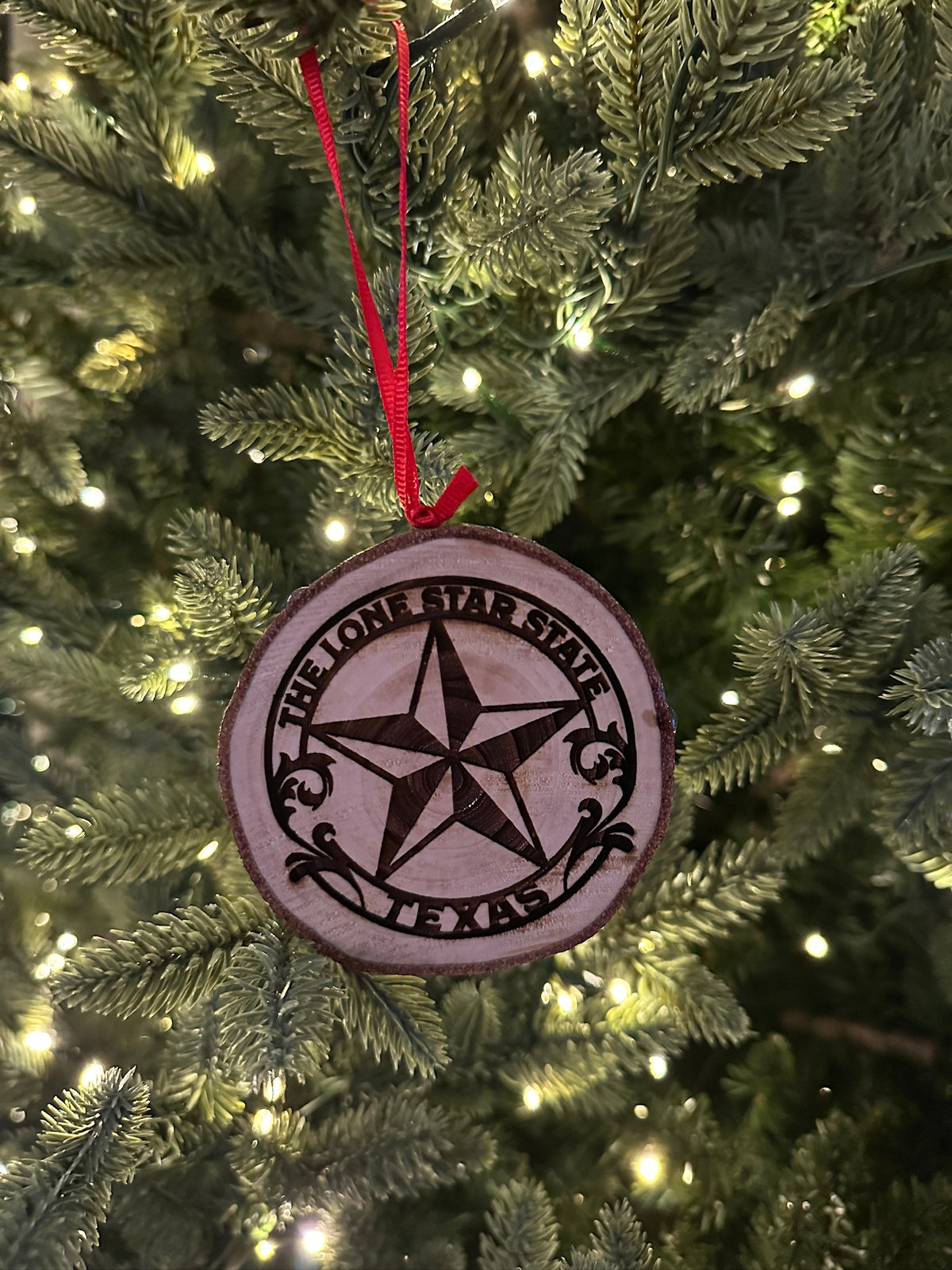 A round wooden Christmas ornament engraved with the Texas state star and 'The Lone Star State of Texas' hanging from a red ribbon on a lit Christmas tree.
