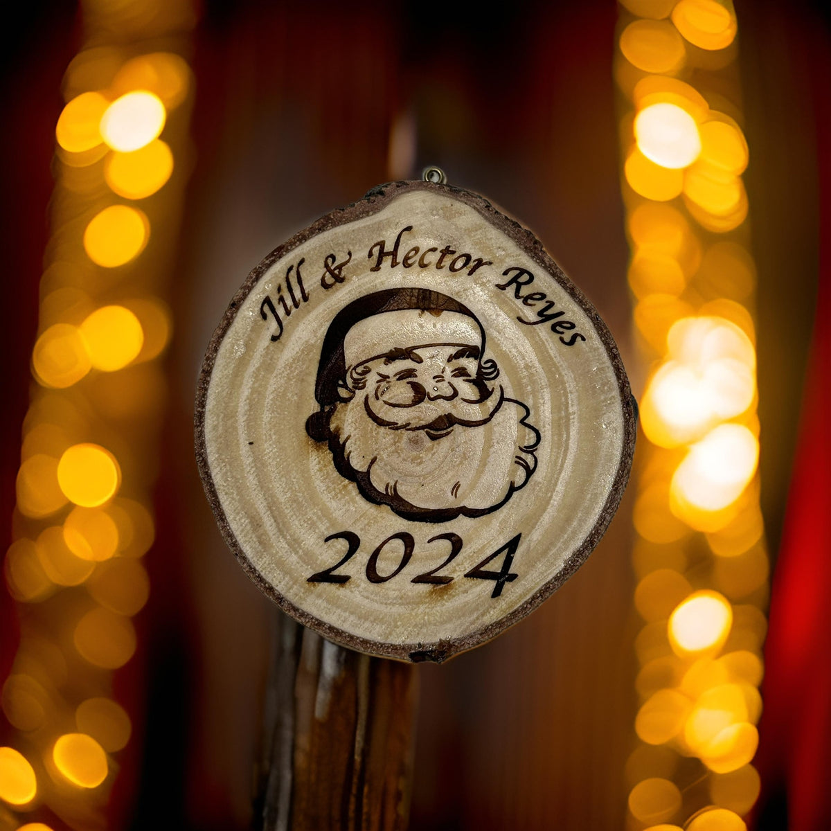 A personalized round wooden Christmas ornament featuring a Santa Claus face and the text 'Jill & Hector Reyes 2024' engraved, shown hanging against a warm, blurred background of golden bokeh lights.