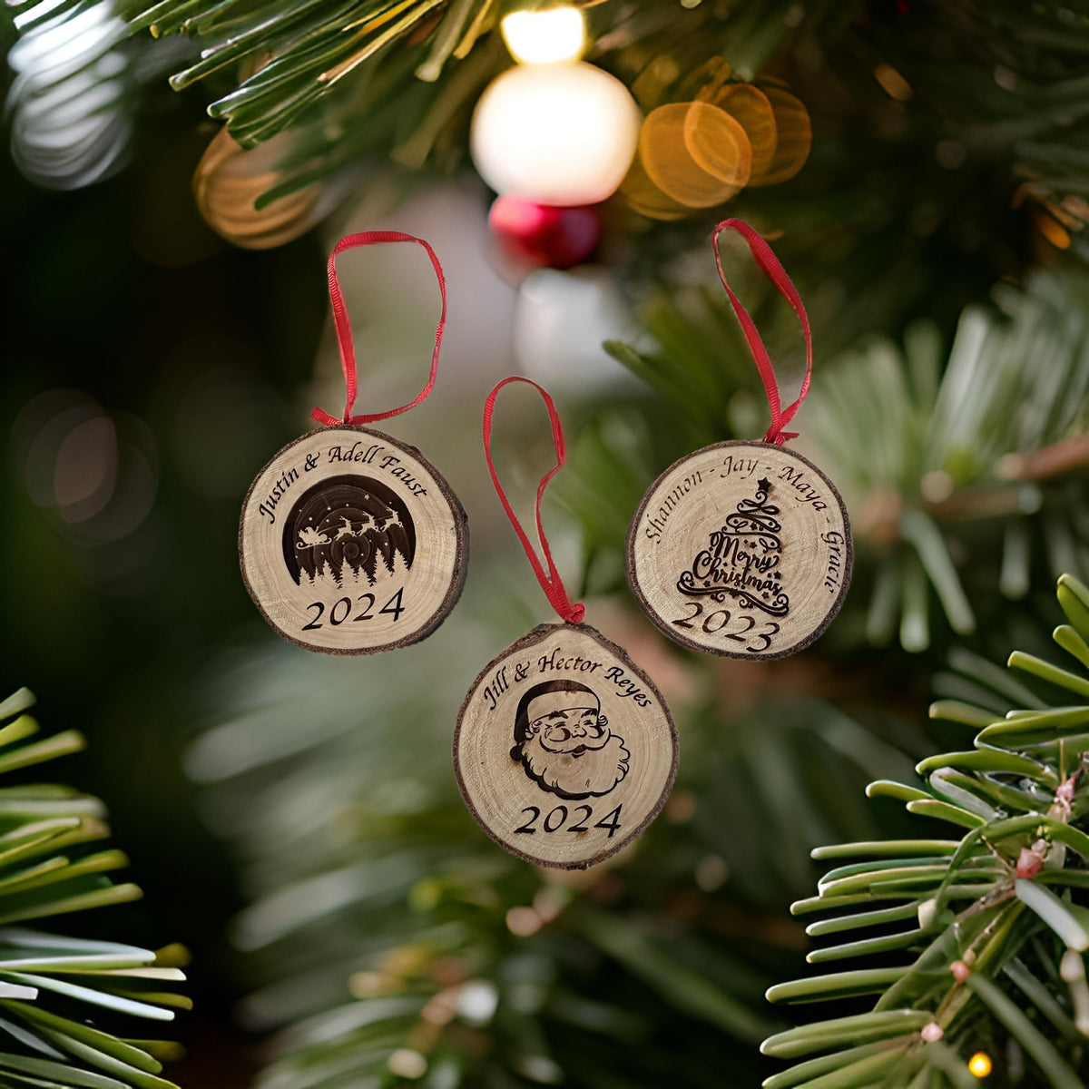 Three personalized wooden Christmas tree ornaments hanging from a pine tree branch with warm holiday lights in the background. The ornaments are engraved with family memories and years 2023 and 2024, each with a red hanging ribbon.