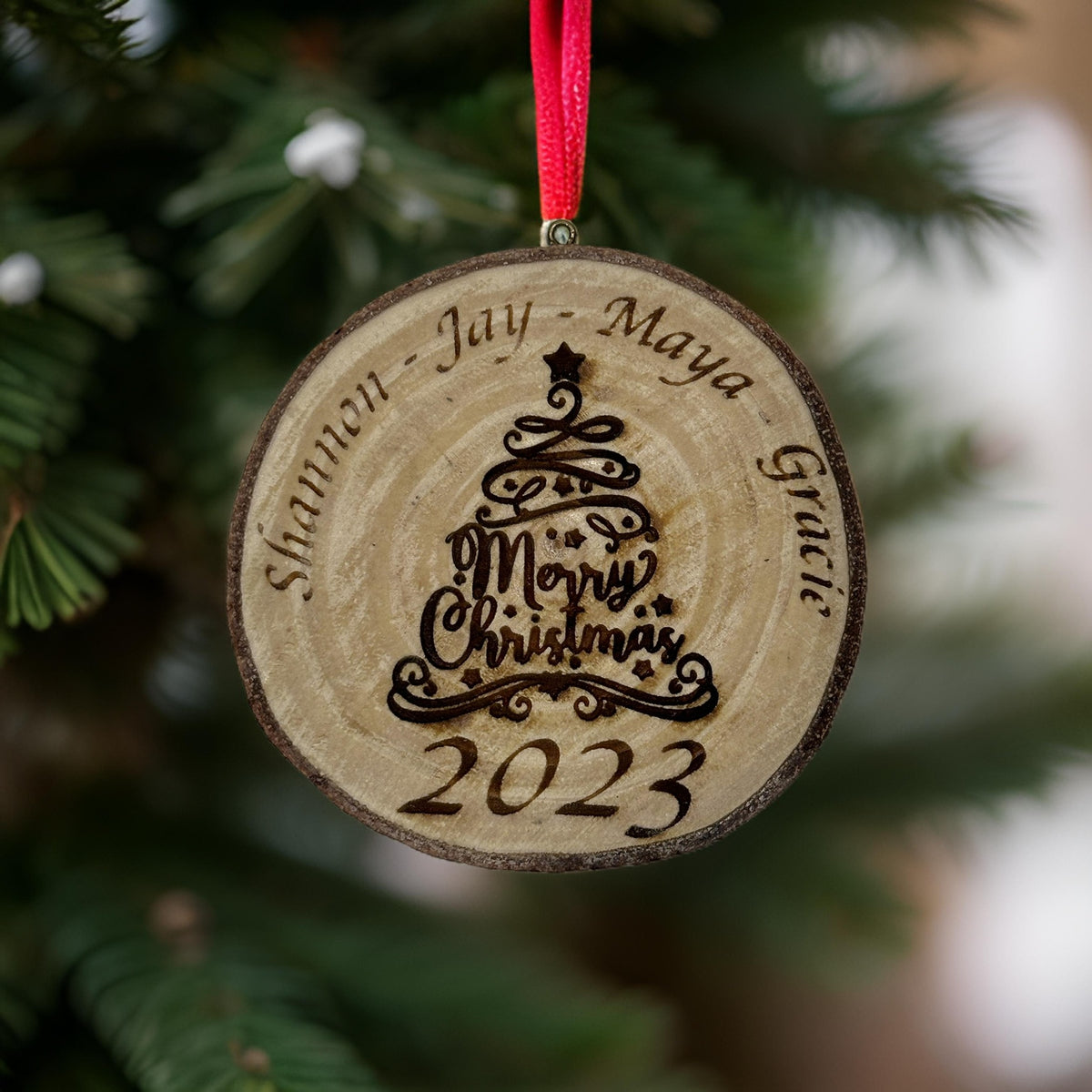A personalized round wooden Christmas ornament hanging on a pine tree. The ornament is engraved with a Christmas tree design, the words 'Merry Christmas 2023', and the names 'Shannon', 'Jay', 'Maya', 'Grace'.