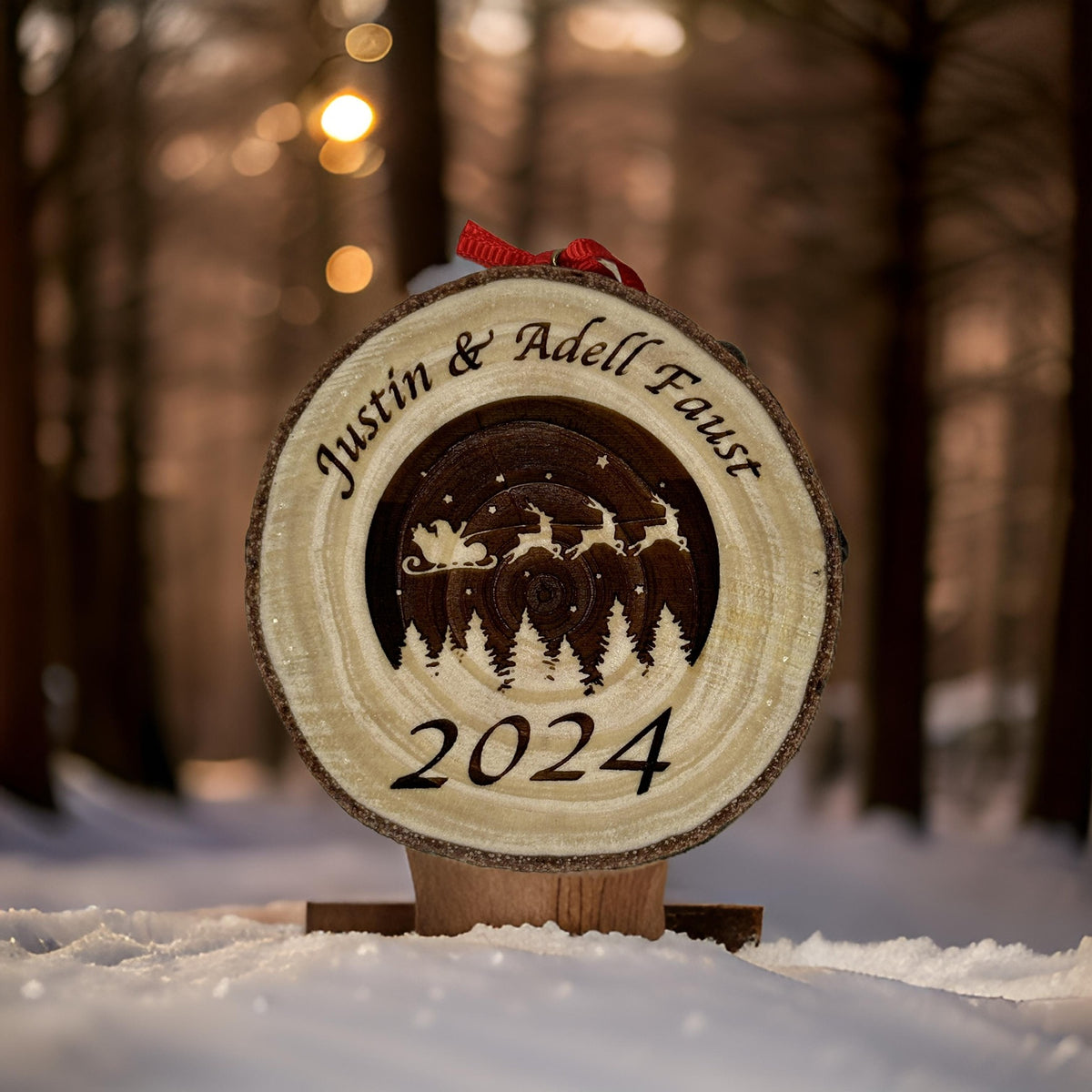 A personalized wooden Christmas ornament carved with a winter forest scene, the names 'Justin & Adell Faust', and '2024', displayed in the snow with a blurred forest background.