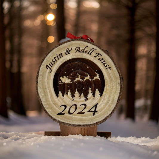 A personalized wooden Christmas ornament carved with a winter forest scene, the names 'Justin & Adell Faust', and '2024', displayed in the snow with a blurred forest background.