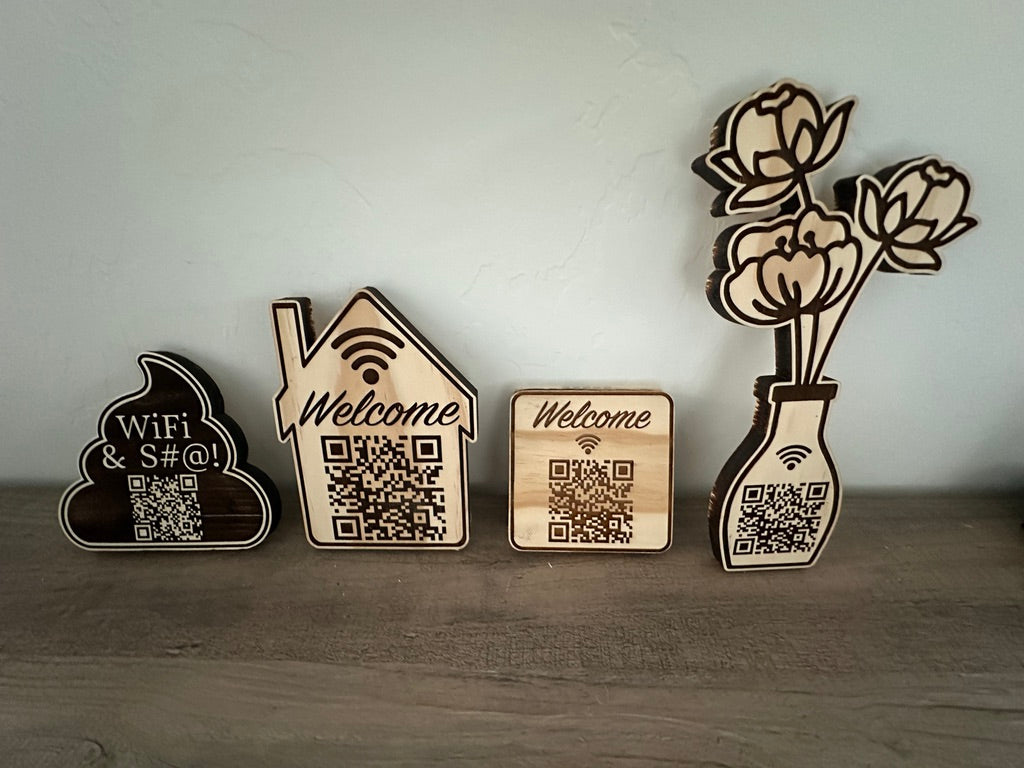 Four laser-cut wooden decorative signs with QR codes and engraved text, displayed on a wooden surface against a light gray wall. The designs include a cloud with 'WiFi & S#@!', a house with 'Welcome', a square wood block with 'Welcome', and a vase with flowers.