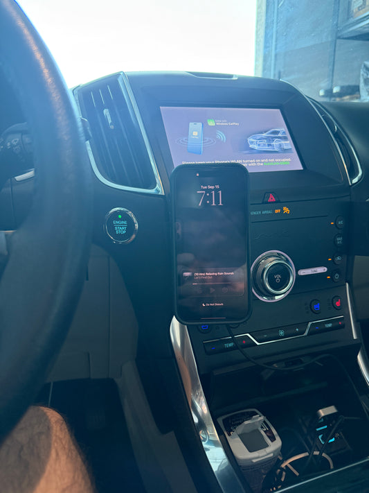 A black smartphone mounted vertically on a dashboard console inside a car, with the infotainment screen and steering wheel visible.