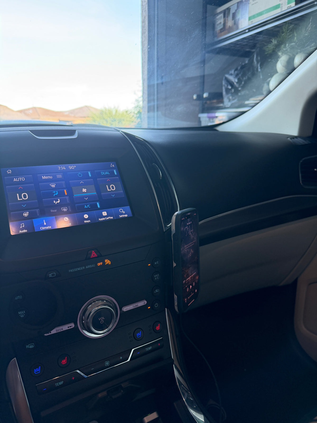 A black smartphone mounted on a holder attached to a car's dashboard console, with the vehicle's infotainment screen and center console visible.