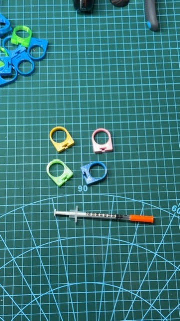 Four small plastic ring-shaped components in yellow, green, pink, and blue are arranged on a green cutting mat alongside a syringe for scale.