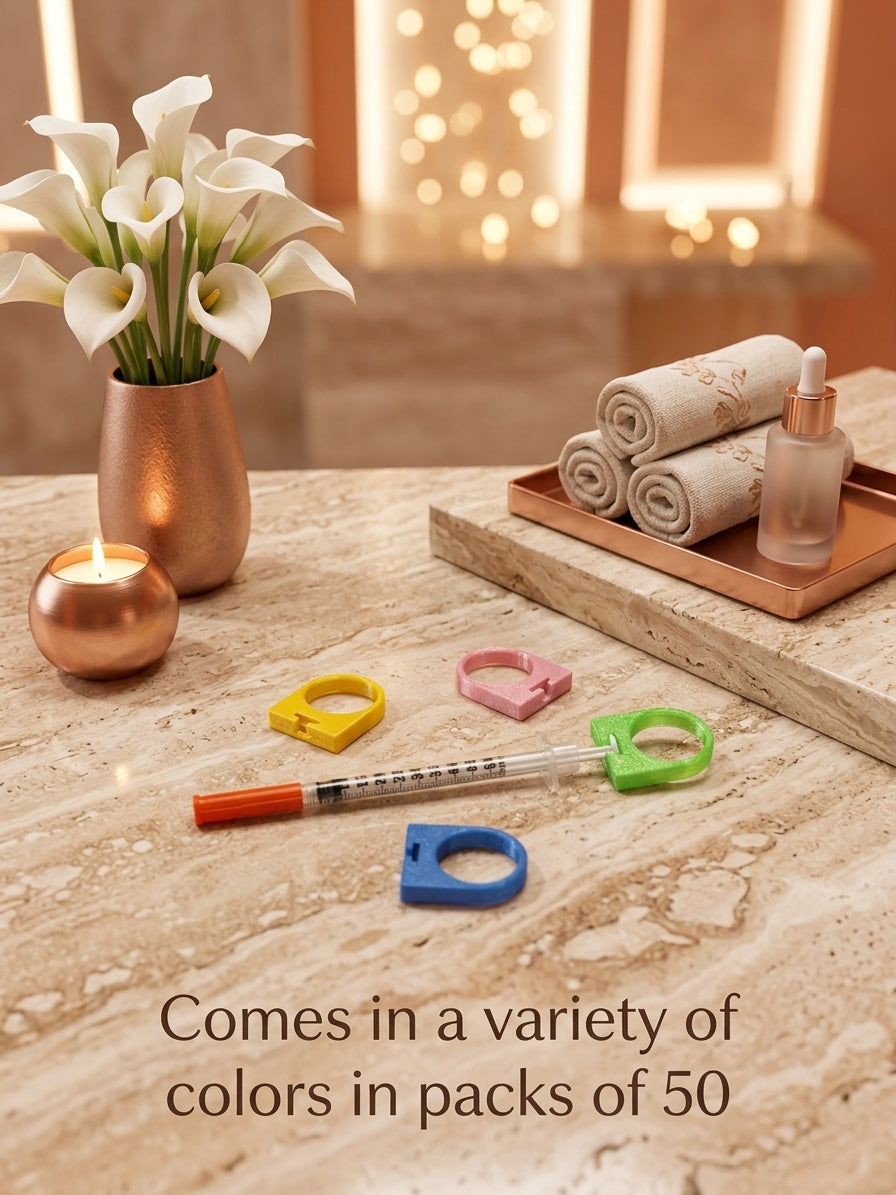 A set of colorful silicone ring-shaped accessories displayed on a marble countertop, alongside a candle, a vase of white calla lilies, rolled towels, and a dropper bottle. Text at the bottom reads, 'Comes in a variety of colors in packs of 50'.