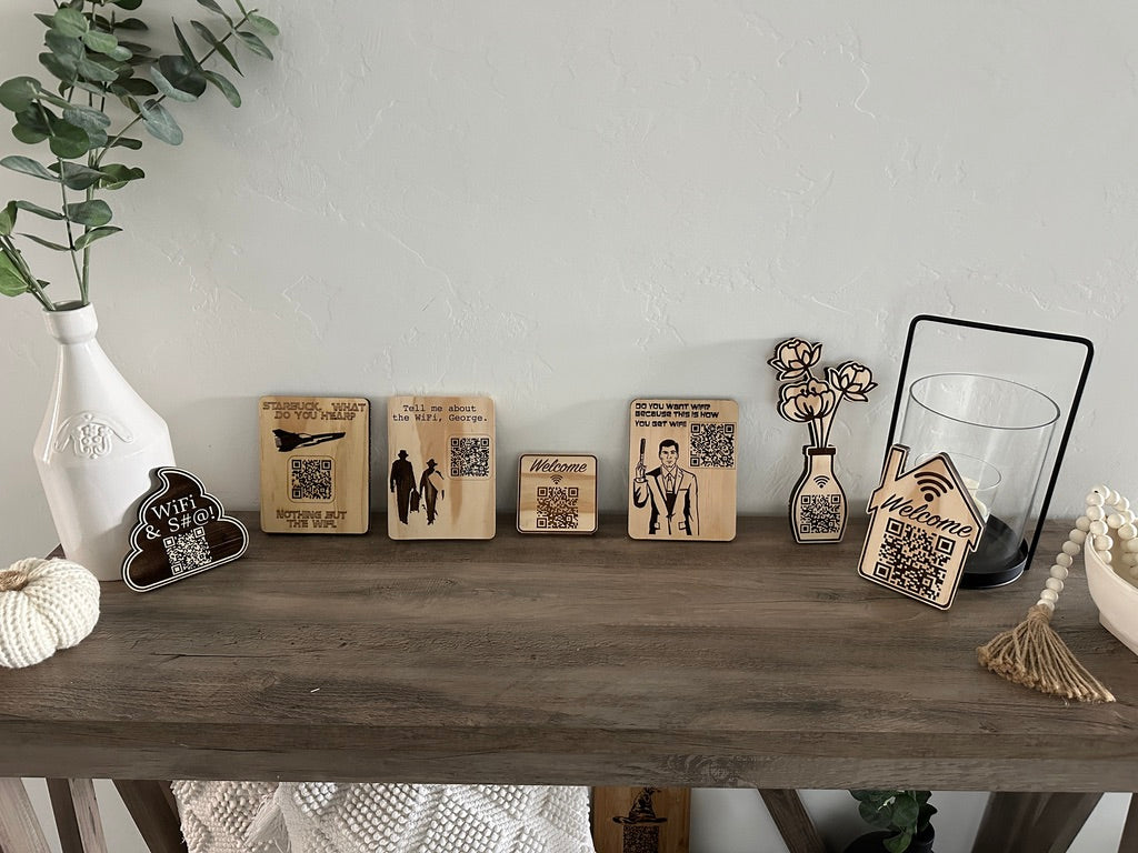 A styled tabletop display featuring several engraved wooden decorative blocks and small vases with printed designs, including QR codes, travel stamps, a couple's silhouette, a man in a suit, a house-shaped sign, and floral motifs, arranged on a rustic wooden console table against a light textured wall.