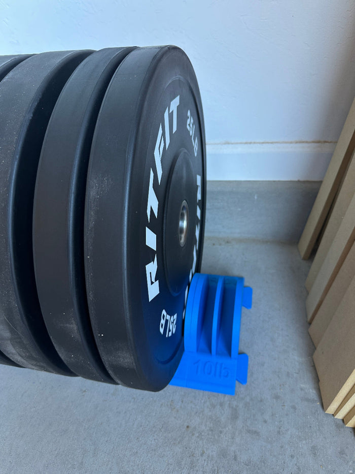 Black bumper plates standing upright in a blue floor holder against a white wall