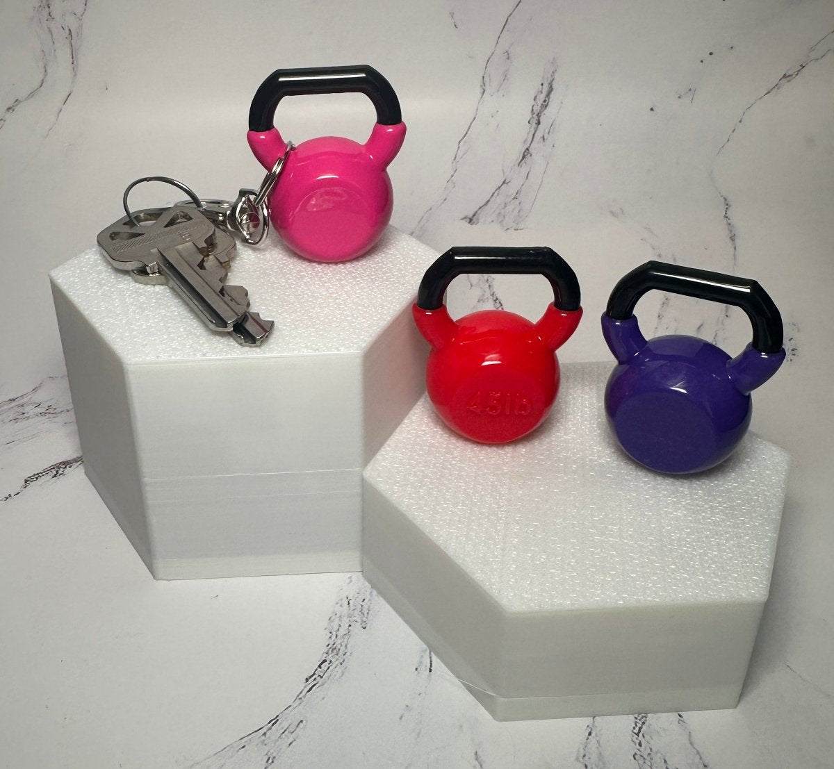 Pink, red and purple mini kettlebell keychains on white stands, one attached to a set of keys
