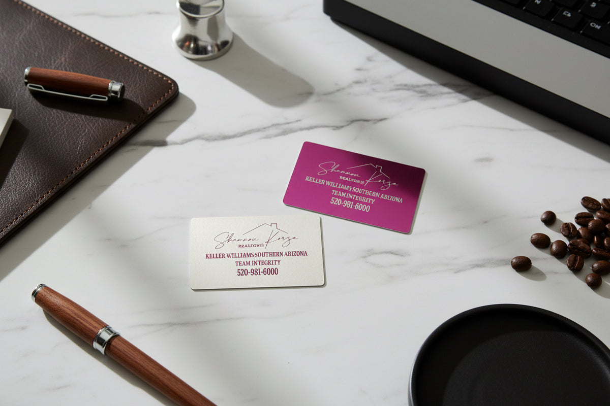 Pink and silver engraved metal business cards on a marble desk with a pen and coffee beans