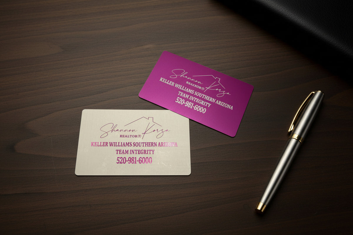 Pink and silver engraved metal business cards on a dark wood desk with a pen