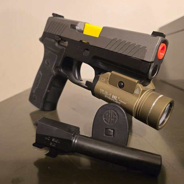 Sig Sauer pistol with a weapon light and yellow chamber flag on a display stand, with a black barrel in front