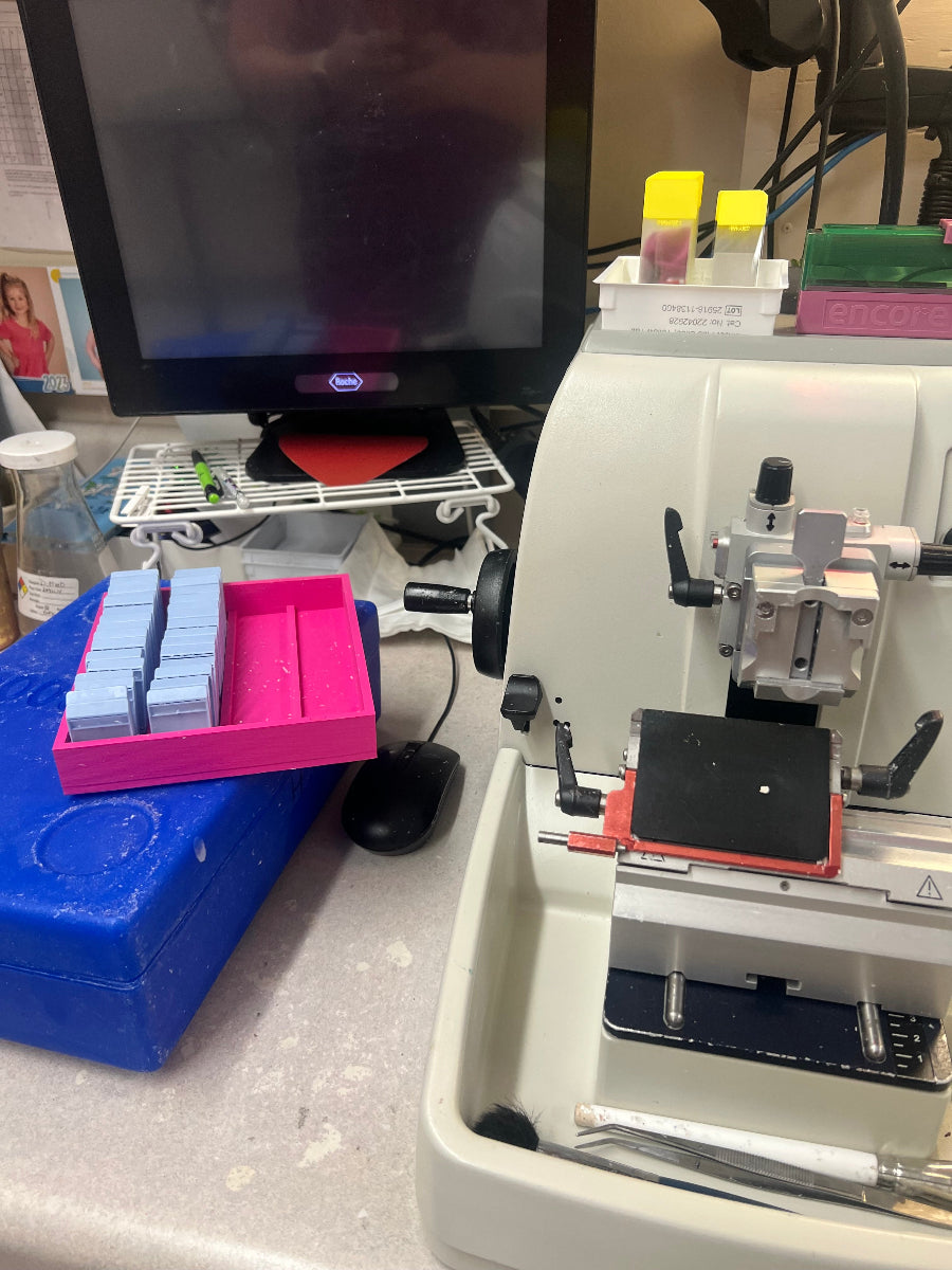 Pink and blue tissue cassette racks holding cassettes next to a microtome in a histology lab