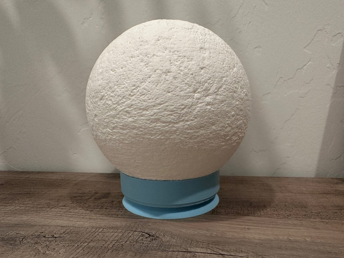 Unlit white moon lamp with crater texture on a blue base on a wood shelf